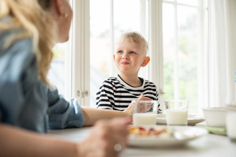  Kuvassa lapsi hymyilee äidilleen ruokapöydän ääressä.