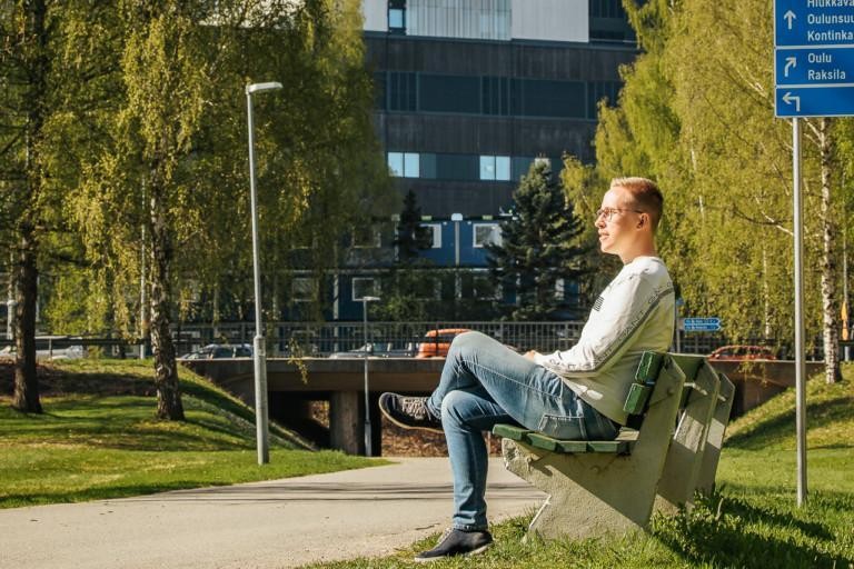  Viljami Kallio istuu puistonpenkillä, taustalla on OYSin uuden sairaalan rakennustyömaa.